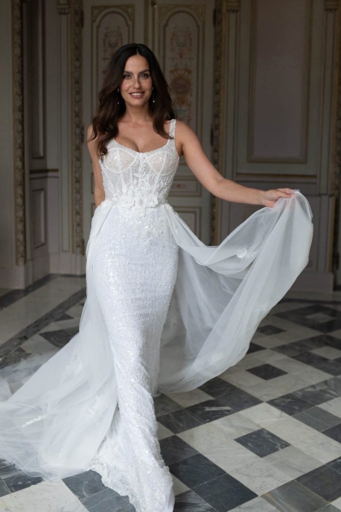 In questa foto, una modella posa nel salone di una dimora storica indossando un abito da sposa a sirena con ricami di paillettes con sopragonna in organza e applicazioni di fiori in vita