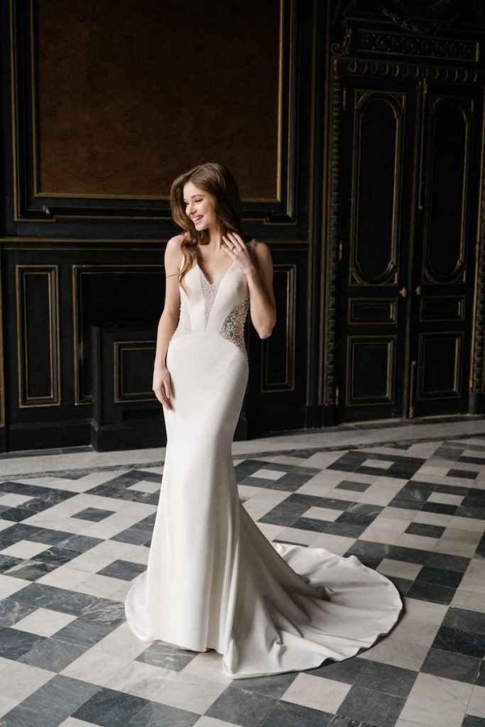 In questa foto, una modella posa nel salone di una dimora storica indossando un abito da sposa a sirena in cady di seta della collezione Abella 2026. Un modello che presenta sui fianchi e sulla scollatura dei dettagli cut out effetto illusion con inserti di perline