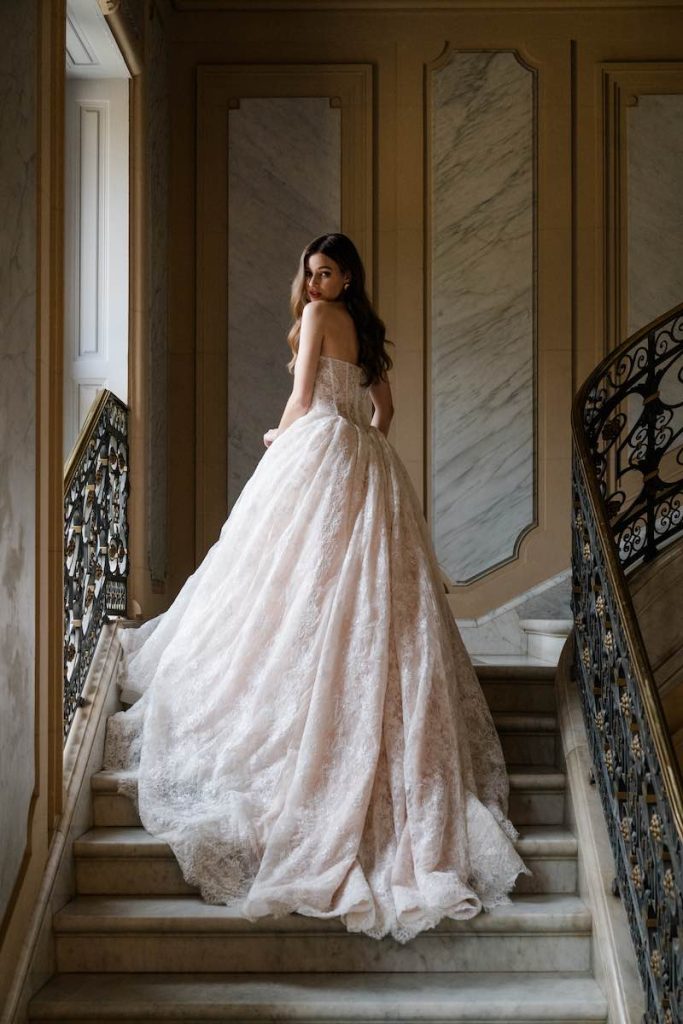 In questa foto, una modella di spalle posa sulla scalinata in marmo di una dimora storica indossando un abito da sposa Abella 2026 dalla silhouette ad A, con bustier a vista, scollo a cuore e in pizzo in nuance di rosa