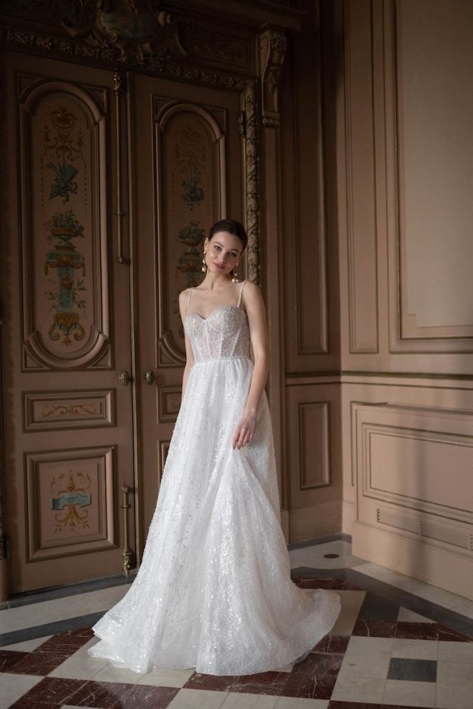In questa foto, una modella posa nel salone di una dimora storica indossando un abito da sposa dalla silhouette ad A, bustier a vista con scollo a cuore e inserti brillanti all over