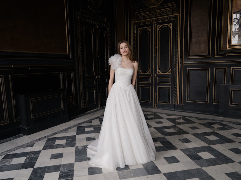 In questa foto, una modella posa nella sala di un salone nobiliare indossando un abito da sposa della collezione Abella 2026, modello scivolato, con un maxi fiore tridimensionale sulla spalla destra