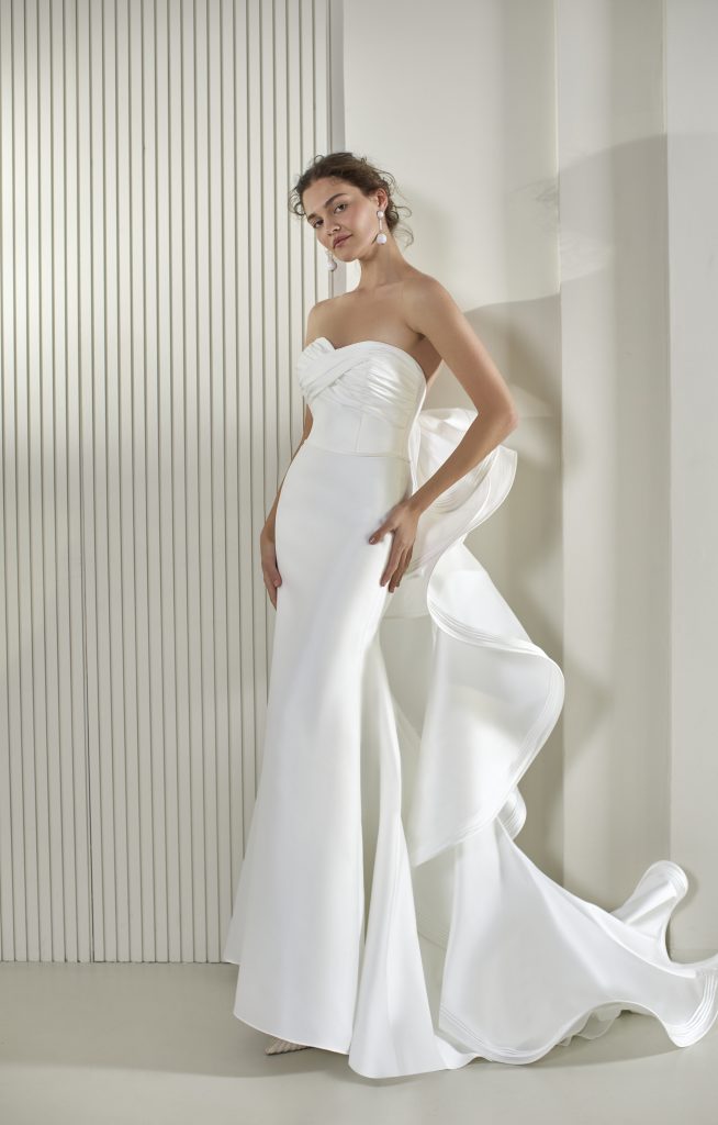 In questa foto, una modella posa in un'elegante stanza dallo stile classico indossando uno degli abiti da sposa a sirena 2026. Il vestito è in pizzo bianco, con profonda scollatura a V, spalline sottili e un taglio che segue le forme del corpo, terminando con un piccolo strascico.