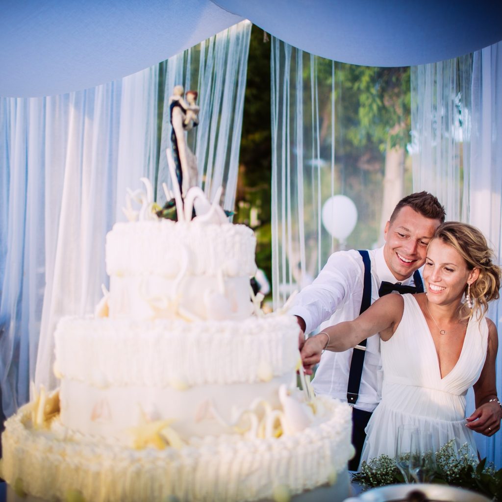 In questa foto si vede una coppia di sposi che taglia una torta nuziale a più piani, decorata con panna e dettagli bianchi. I due sono sorridenti, vestiti in abiti eleganti, sotto un gazebo con tendaggi chiari. L’atmosfera è romantica e serena.