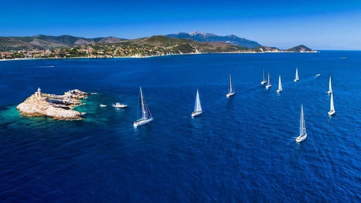In questa foto si vede una fila ordinata di barche a vela che solcano un mare blu intenso, costeggiando un’isoletta con un piccolo faro. Sullo sfondo, la costa verde dell’Isola d’Elba si estende fino alle montagne. La scena comunica libertà e armonia con la natura.