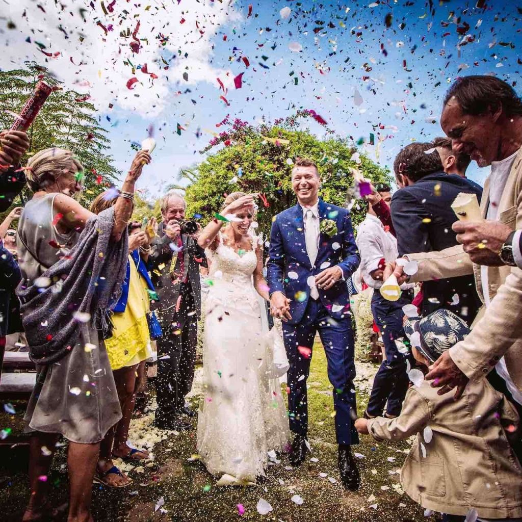 In questa foto si vede una coppia di sposi al centro dell’inquadratura, avvolta da una pioggia colorata di coriandoli, circondata da invitati festanti che applaudono e scattano foto. La scena è all’aperto, illuminata dalla luce del giorno e piena di gioia.