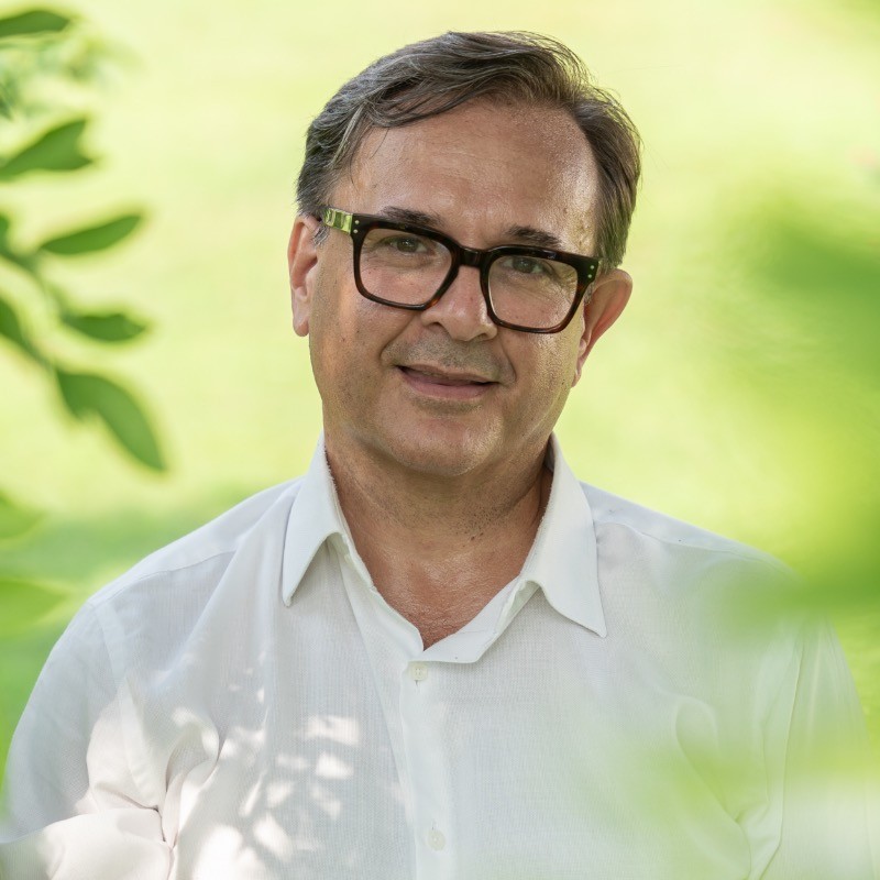 In questa foto si vede l'architetto Angelo Garini, direttore del progetto, che si presenta come un uomo maturo con occhiali e camicia bianca, sorridente, seduto in un ambiente naturale con sfondo verde sfocato. La luce è soffusa e naturale, creando un’atmosfera intima e accogliente.