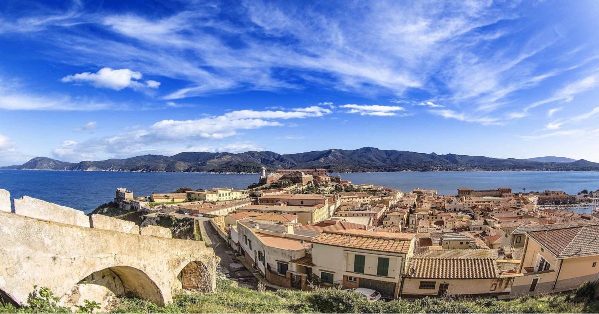 In questa foto si vede una panoramica mozzafiato dell’Isola d’Elba con un cielo azzurro ricco di nuvole bianche e leggere. In primo piano i tetti delle case color ocra e le mura antiche, sullo sfondo il mare blu e le montagne dell’isola.
