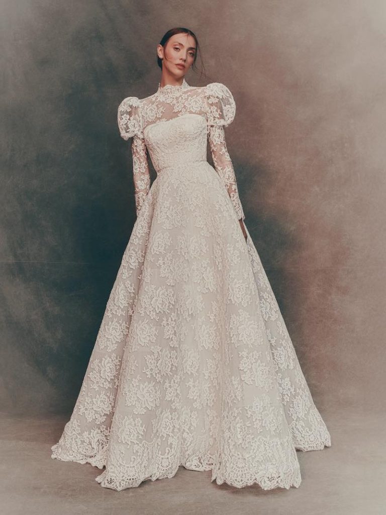 In questa foto una modella posa indossando un abito trasformabile di Atelier Pronovias 2026 ad A in pizzo floreale, maniche lunghe e maniche balloon 