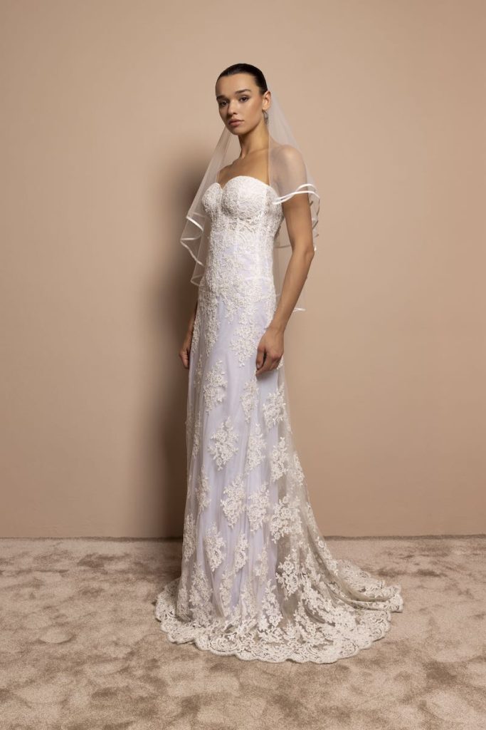 In questa foto, una modella posa indossando un abito da sposa 2026 a sirena in pizzo con bustier steccato e scollo a cuore. La modella indossa anche un velo corto con bordo in raso