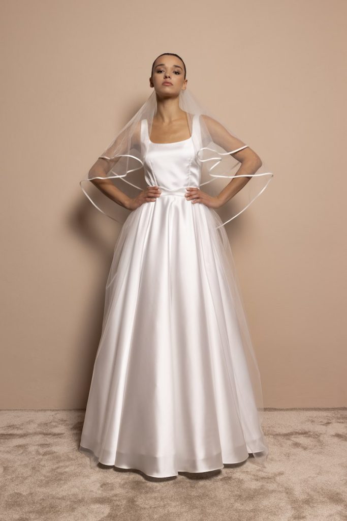 In questa foto, una modella posa indossando un abito da sposa Daniela de Souza 2026  in mikado di seta con silhouette ad A con scollatura quadrata. La modella indossa anche un velo corto con bordo di raso
