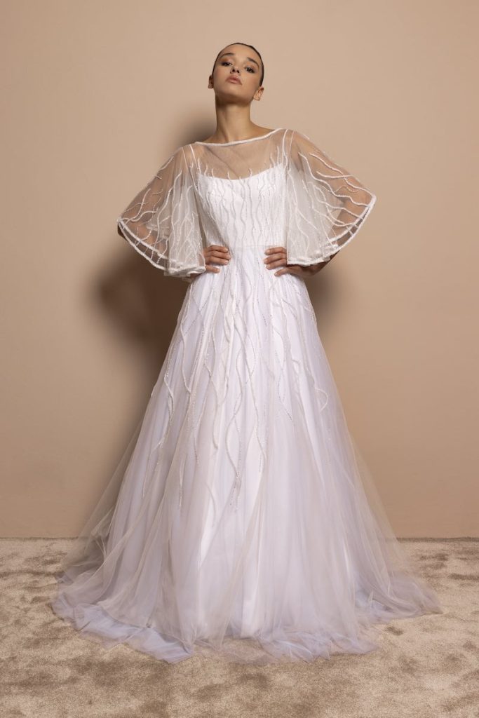 In questa foto, una modella indossa un abito da sposa 2026 di Daniela De Souza in tulle con riflessi di colore argento, con silhouette ad A. La modella indossa anche una cappa con maniche a corolla 