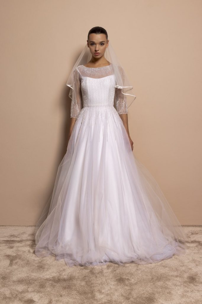 In questa foto, una modella indossa un abito da sposa 2026 di Daniela De Souza in tulle con riflessi di colore argento, con silhouette ad A, maniche a tre quarti, girocollo 