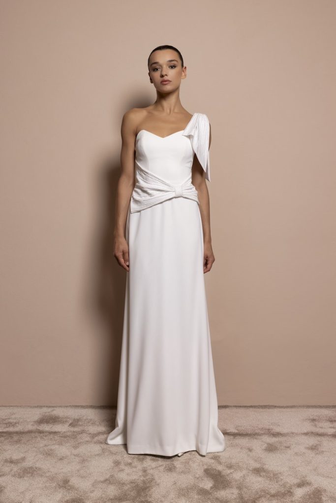 In questa foto, una modella indossa un abito da sposa scivolato in cady di seta, monospalla con fiocco sulla spalla e intreccio sul punto vita