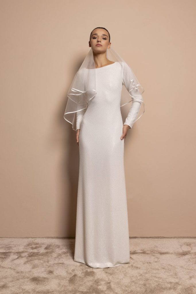 In questa foto, una modella indossa un abito da sposa a sirena con ricami di paillettes, maniche lunghe, scollo ovale. Indossa anche un velo corto con bordo in raso