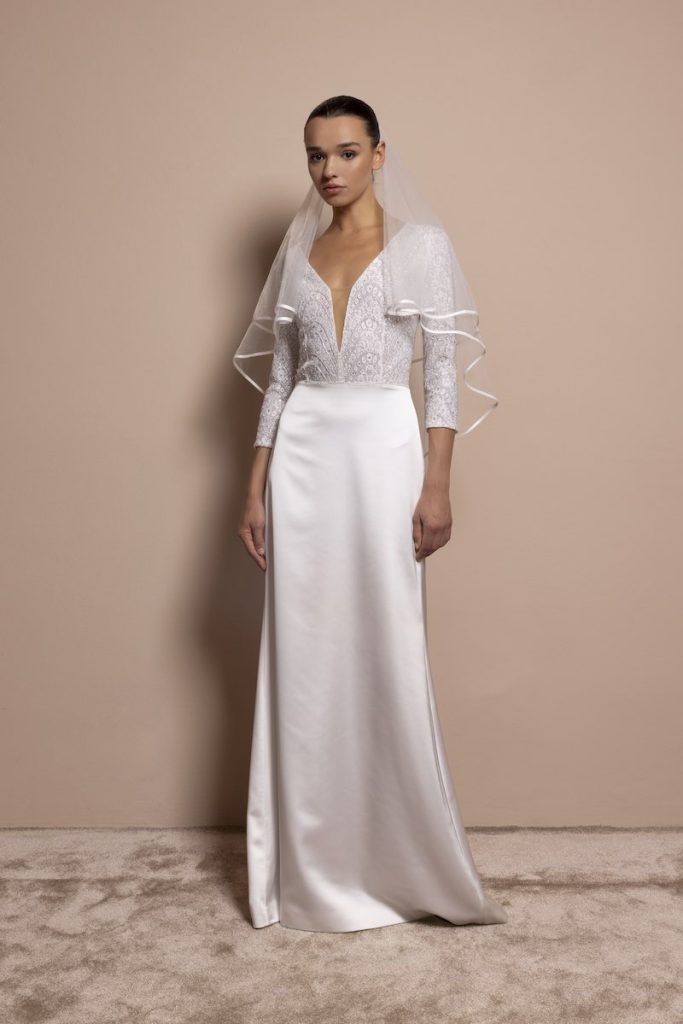 In questa foto, una modella indossa un abito da sposa a sirena con gonna in mikado di seta, corpetto in pizzo ricamato con maniche a tre quarti e scollo a V profondo