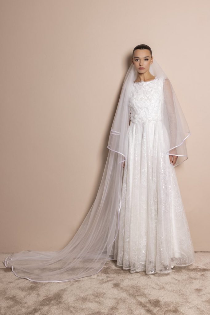 In questa foto, una modella indossa un abito da sposa 2026 di Daniela De Souza dalla silhouette scivolata con petali 3D sul corpetto e riflessi luminosi di paillettes sulla gonna. La modella indossa anche anche un velo con bordo di raso