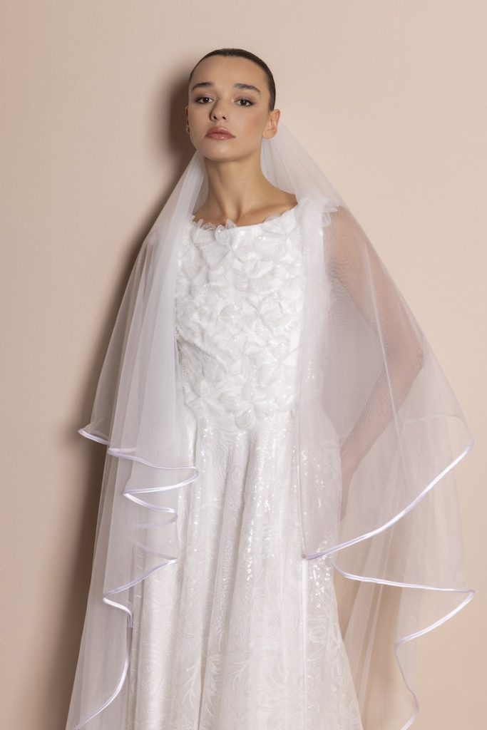 In questa foto, una modella inquadrata a mezzo busto che indossa un abito da sposa 2026 di Daniela De Souza dalla silhouette scivolata con petali 3D sul corpetto e riflessi luminosi di paillettes sulla gonna. La modella indossa anche anche un velo con bordo di raso