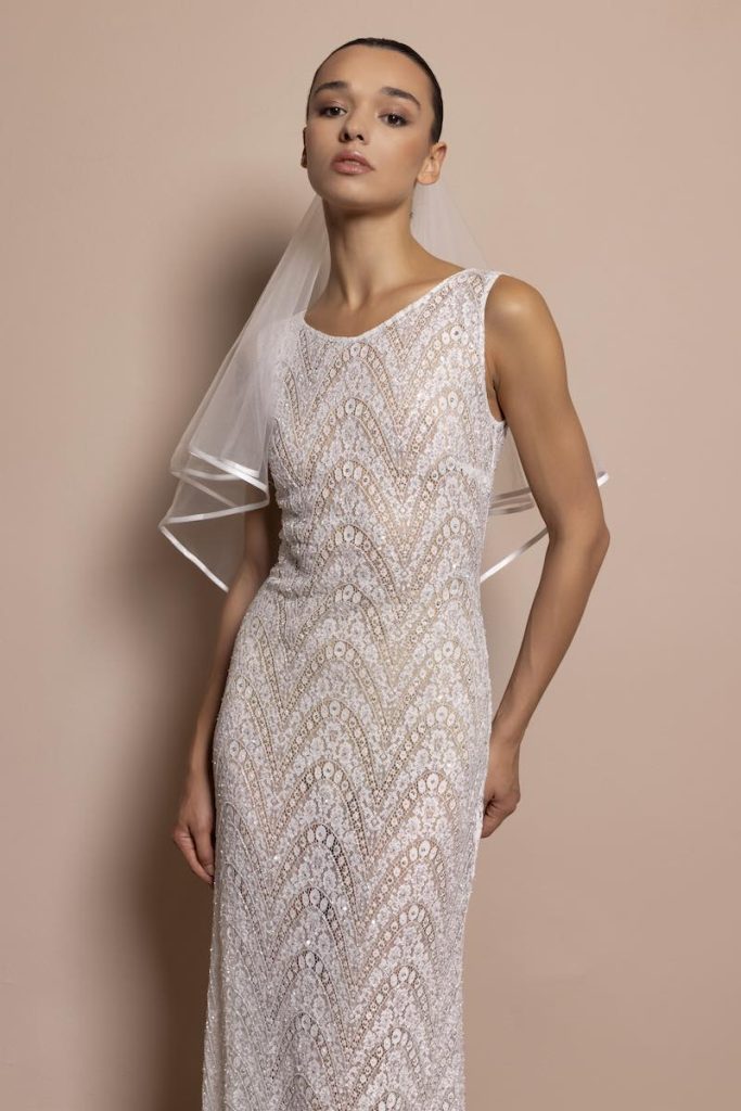 In questa foto, una modella indossa uno degli abiti da sposa Daniela De Souza 2026 con silhouette a sirena in pizzo con ricami preziosi abbinato ad un velo corto con bordo in raso 