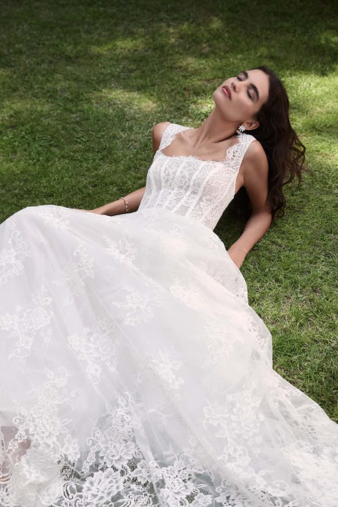 In questa foto, una modella sdraiata sul prato indossa Beatrice, uno degli abiti da sposa Atelier Emé 2026 dalla silhouette ad A, in pizzo Chantilly, con corpetto steccato, scollo quadrato e manichette in pizzo