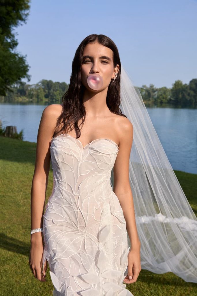 In questa foto, una modella posa su un prato a bordo lago mentre indossa Gea, uno degli abiti da sposa Atelier Emé 2026 con scollo a cuore ed effetto origami e lungo velo con ricami floreali