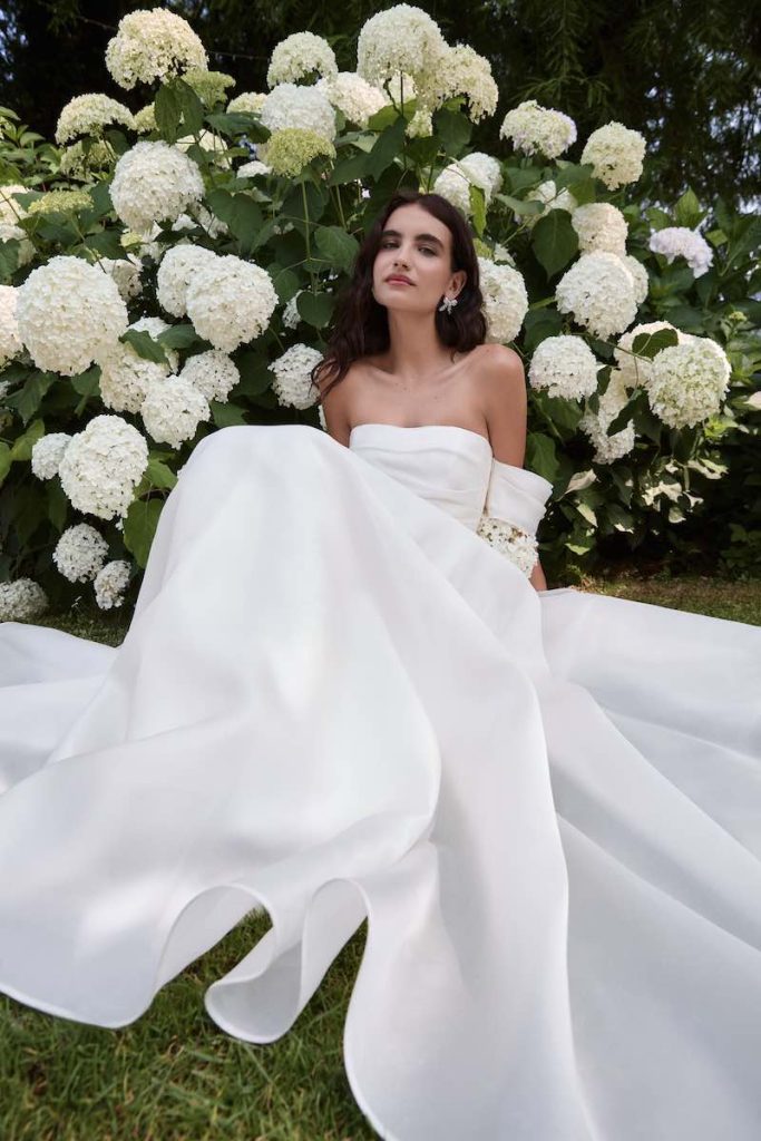 In questa foto, una modella sdraiata sul prato tra ortensie di colore bianco indossa Luana, uno degli abiti da sposa Atelier Emé 2026 caratterizzato da un ampia gonna in mikado luminoso