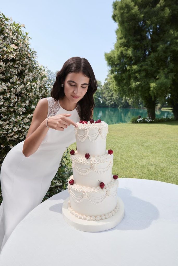 In questa foto, una modella tocca con un dito per assaggiarla una torta nuziale mentre indossa Marina, uno degli abiti da sposa Atelier Emé 2026 dalla silhouette a sirena in cady con scollatura girocollo e maniche in pizzo