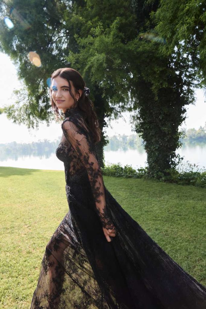 In questa foto, una modella posa camminando su un prato e indossando l'abito da sposa Samira, in pizzo di colore nero, con maniche lunghe e lungo strascico 
