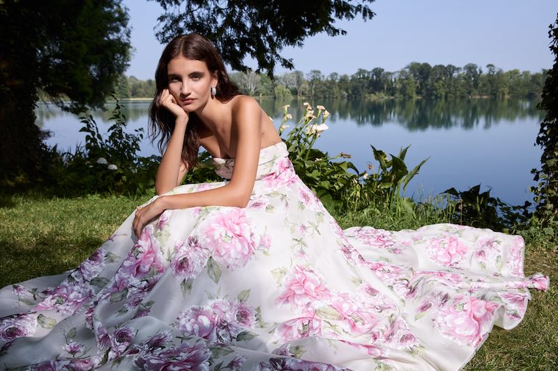 In questa foto, una modella posa seduta su un prato indossando l'abito da sposa Susan, dalla silhouette ampia e in tessuto dipinto con fiori di colore rosa e bianco