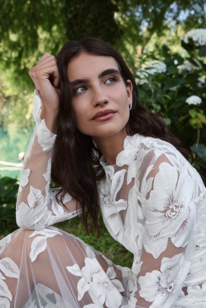 In questa foto, il primo piano di una modella che posa su un prato, seduta, indossando Tessa, uno degli abiti da sposa Atelier Emé 2026 con maniche lunghe e tessuto floreale effetto tattoo su tutto il corpo