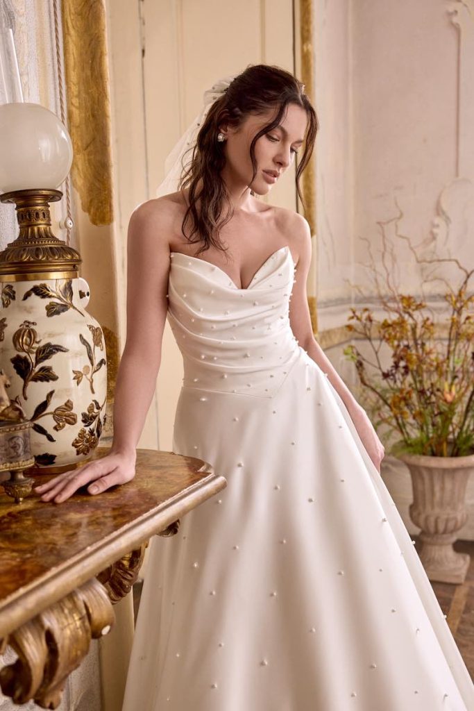 In questa foto, una modella posa indossando un abito da sposa Justin Alexander SS 2026 modello ad A in mikado con drappeggi sul corpetto e inserti di perline all over 