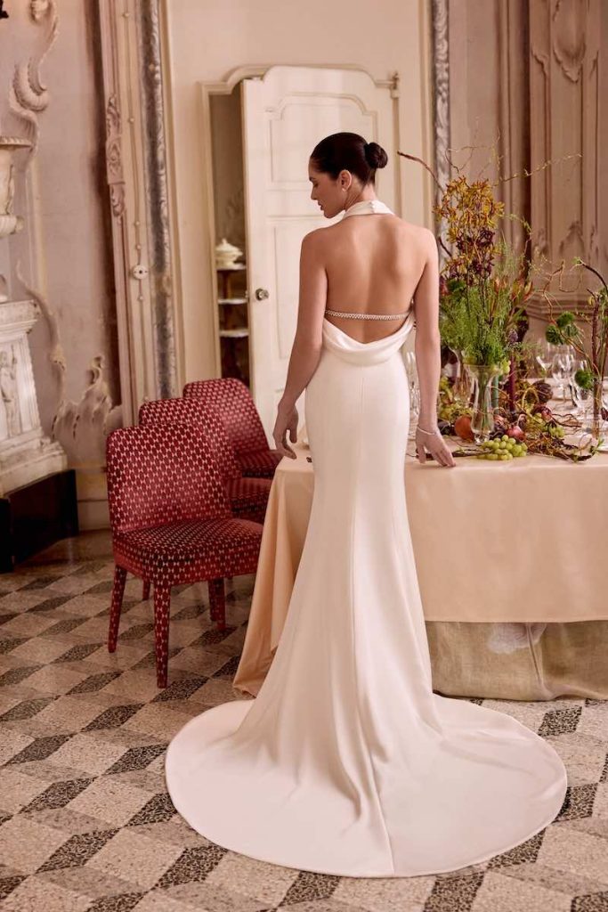 In questa foto, una modella posa di spalle indossando un abito da sposa Justin Alexander SS 2026 a sirena con schiena scoperta e dettaglio gioiello