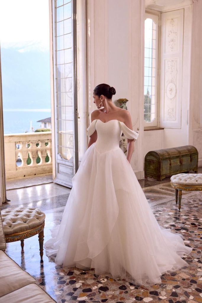 In questa foto, una modella posa indossando uno degli abiti da sposa Justin Alexander SS 2026 modello ad A in tulle e organza con scollo off shoulder e busto a vita basca 