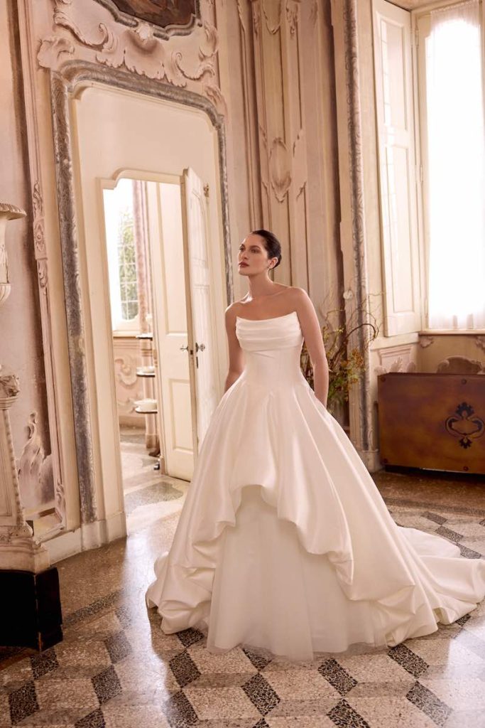In questa foto, una modella posa indossando uno degli abiti da sposa Justin Alexander SS 2026 modello ad A in mikado e tulle con drappeggio sul busto e ruches sulle lunghezze
