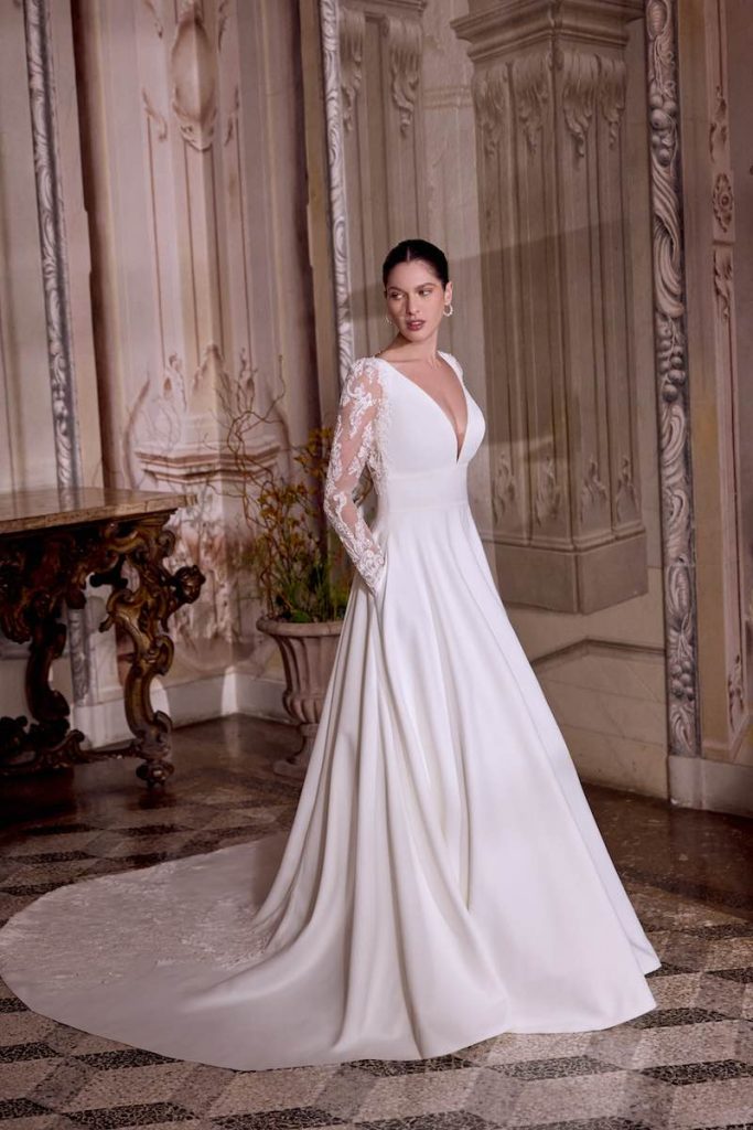 In questa foto, una modella posa indossando uno degli abiti da sposa Justin Alexander SS 2026 modello ad A con scollo a V, maniche lunghe in pizzo floreale illusion e lungo strascico