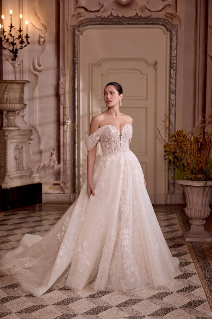 In questa foto, una modella posa indossando uno degli abiti da sposa Justin Alexander SS 2026 modello ad A, in tulle glitter con inserti floreali preziosi con scollatura off shoulder