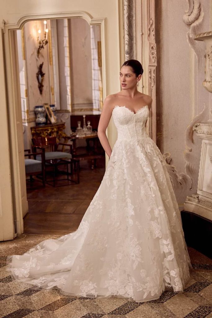In questa foto, una modella posa indossando uno degli abiti da sposa Justin Alexander SS 2026 modello ad A in pizzo floreale con scollo a cuore e bustier steccato 