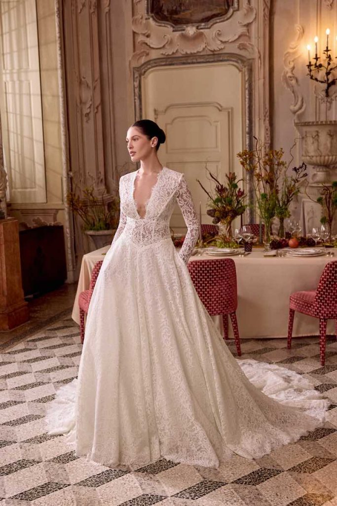 In questa foto, una modella posa indossando uno degli abiti da sposa Justin Alexander SS 2026 modello ad A in pizzo con scollo a V profondo e maniche lunghe 