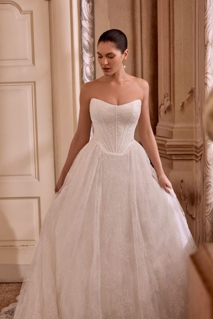 In questa foto, una modella posa indossando uno degli abiti da sposa Justin Alexander SS 2026 modello ad A con scollo dritta e bustier steccato a vita basca