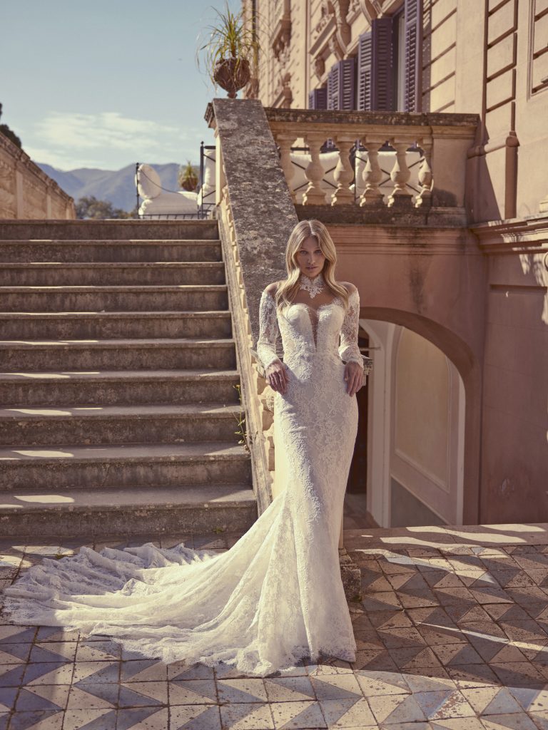 In questa foto, una sposa si trova all’aperto davanti a una scalinata in pietra, indossando uno degli abiti da sposa a sirena 2026. Il vestito è in pizzo con maniche lunghe e trasparenti, scollatura profonda a cuore e un lungo strascico che si apre sul pavimento alle sue spalle.