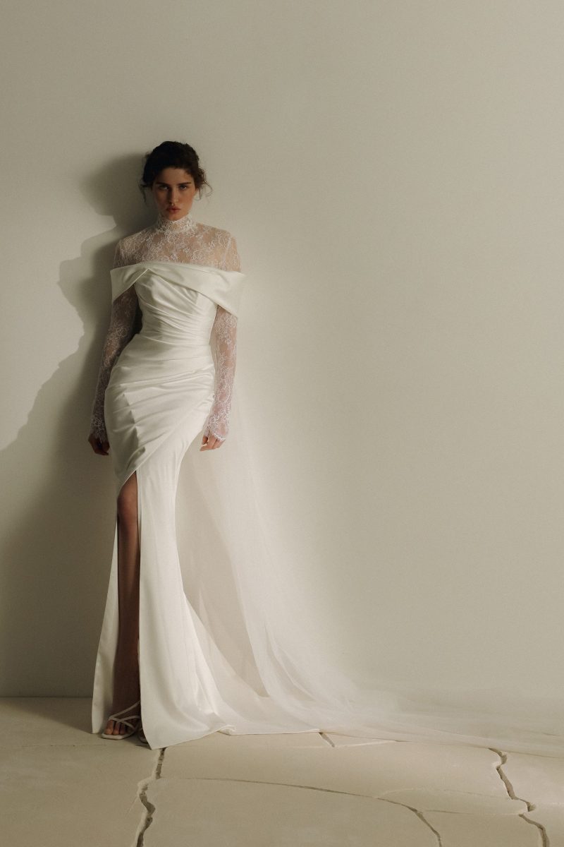 In questa foto, una modella posa con uno degli abiti da sposa a sirena 2026 della maison Eva Lendel. Il vestito, raffinato e moderno, ha una struttura drappeggiata, maniche lunghe in pizzo trasparente e un corpetto che lascia le spalle scoperte, con un lungo strascico di tulle sul retro