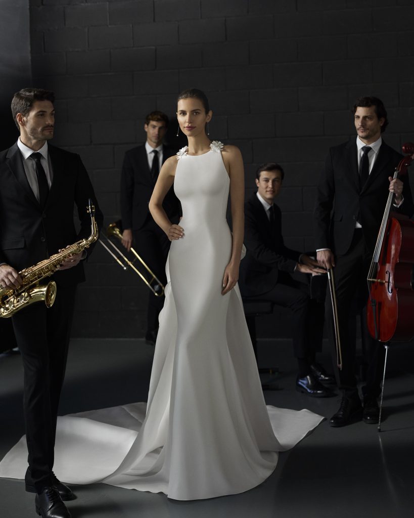 In questa foto, una modella si trova in uno studio elegante circondata da musicisti. Indossa uno degli abiti da sposa a sirena 2026 con un design minimalista e moderno, in tessuto liscio e bianco, caratterizzato da una scollatura all’americana con applicazioni floreali sulle spalle e un lungo strascico posteriore.