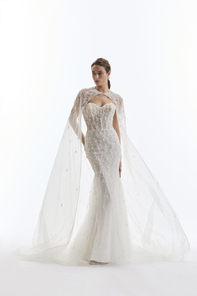 In questa foto, una modella sfoggia un elegante esempio di abiti da sposa a sirena 2026 della collezione Demetrios. Il vestito è interamente decorato con perline e paillettes e presenta un corpetto semi-trasparente a cuore, completato da un mantello in tulle ricamato che crea un effetto etereo.