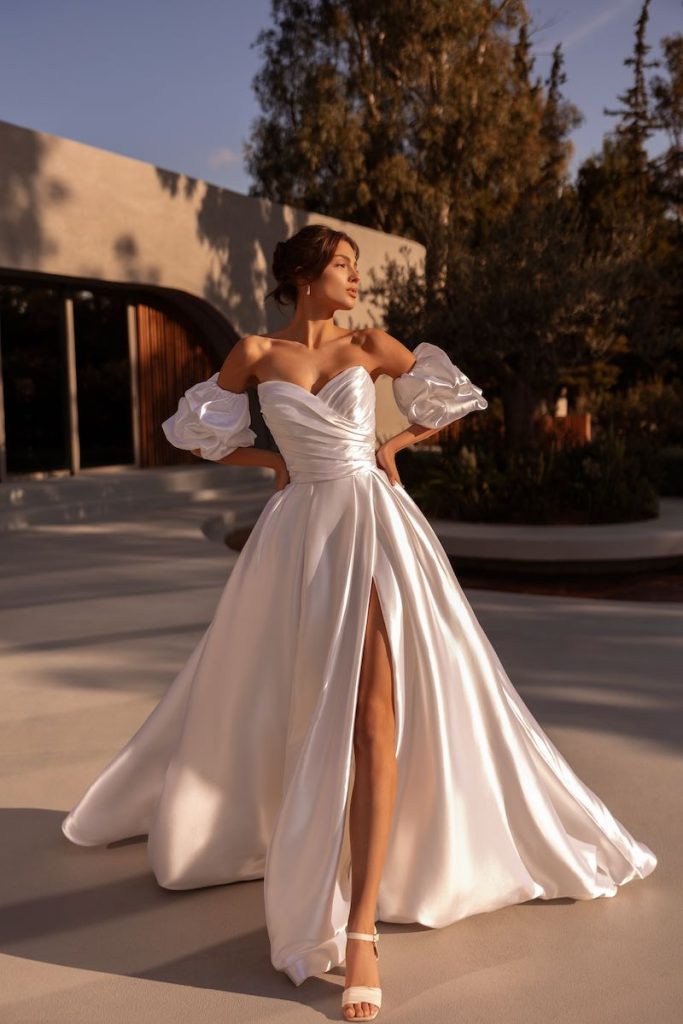 In questa foto, una modella posa indossando un abito della collezione sposa Demetrios 2026 in mikado luminoso, con silhouette ad A, spacco frontale e due manicotti balloon staccabili