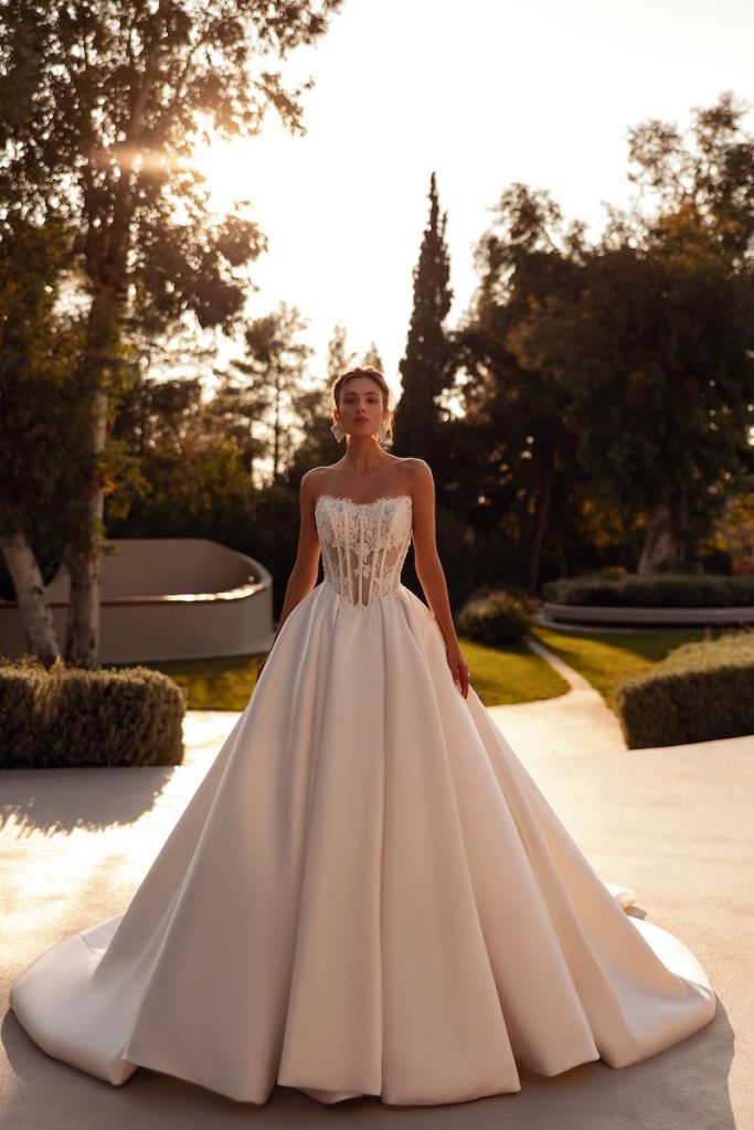 In questa foto, una modella posa indossando un abito della collezione sposa Demetrios 2026 dalla silhouette principessa con gonna in mikado voluminoso e bustier steccato a vista con ricami di pizzo