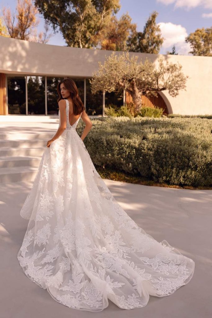 In questa foto, una modella posa di spalle indossando un abito della collezione sposa Demetrios 2026 con tulle ricamato con intarsi floreali e una lunga coda