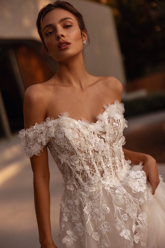 In questa foto, una modella inquadrata a mezzo busto posa mostrano il corpetto con scollo omerale di un abito da sposa Demetrios con applicazione di fiori 3D