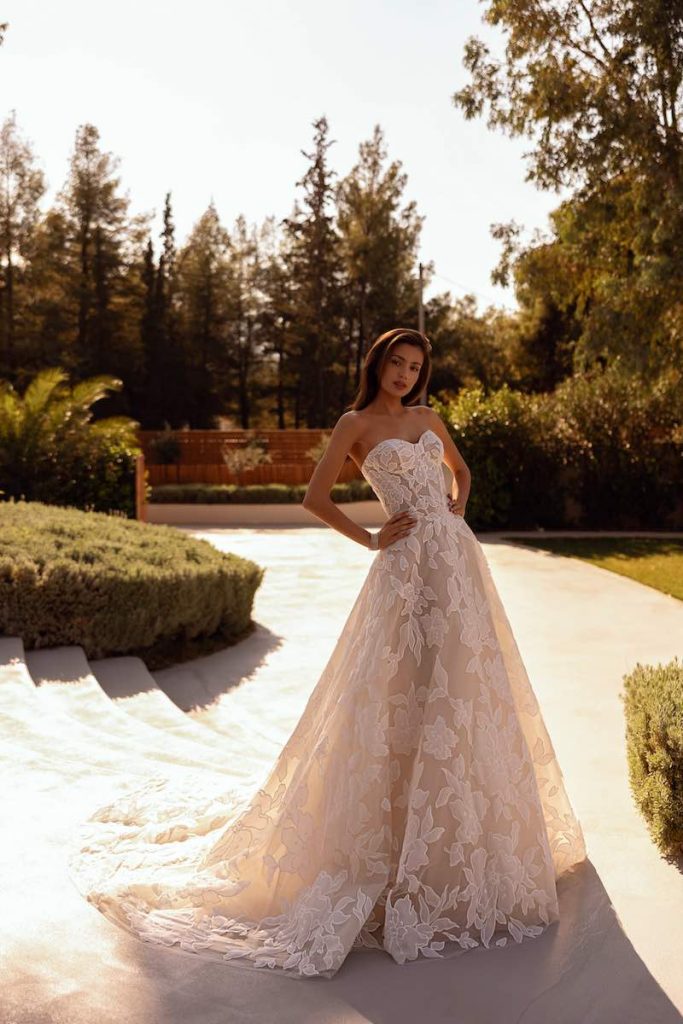 In questa foto, una modella posa indossando un abito della collezione sposa Demetrios 2026 con silhouette ad A, corpetto bustier e ricami floreali all over