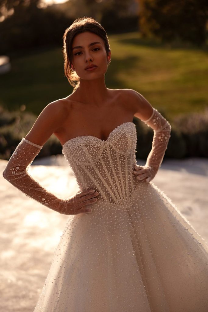 In questa foto, una modella posa indossando un abito della collezione sposa Demetrios 2026 con corpetto steccato a vista con scollo a cuore  ricamato con perline 