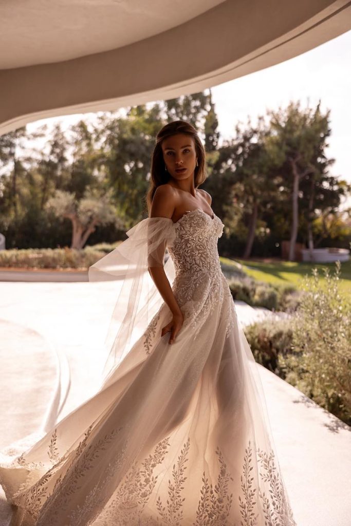 In questa foto, una modella posa indossando un abito della collezione sposa Demetrios 2026 dalla linea ad A in organza con ricami di fiori e foglie e maniche staccabili