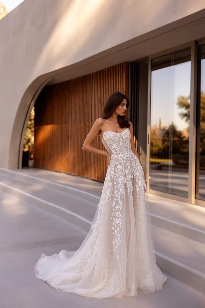 In questa foto, una modella posa indossando un abito della collezione sposa Demetrios 2026 dalla linea scivolata, in tulle glitter con corpetto bustier e ramage floreali che dal corpetto scende sulle lunghezze 